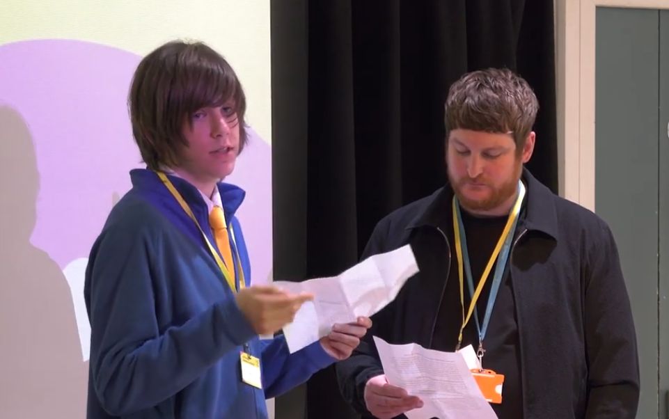 A person holding a piece of paper making a speech stood beside another person